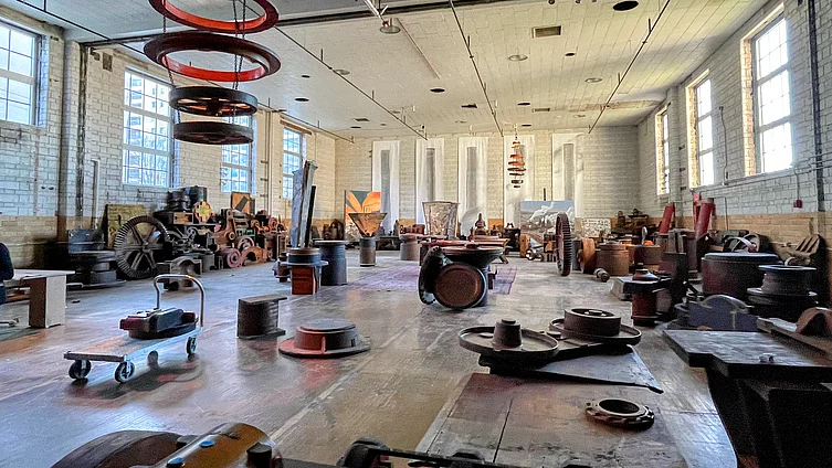Spacious industrial warehouse interior with hanging red rings, scattered machinery, gears, wheels, barrels on wooden floor and white brick walls. 