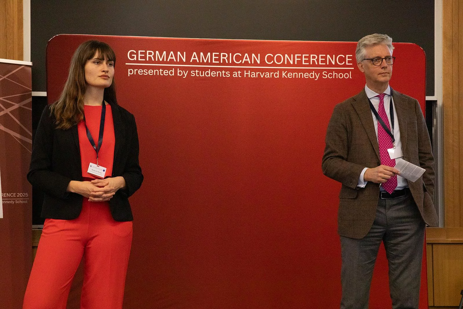 Marlies Murray (Program Officer for Domestic Political Affairs) in red dress and black cardigan stands beside Dr. Reinhard Krumm (Representative to the U.S. and Canada) in tweed jacket before red German American Conference banner. 