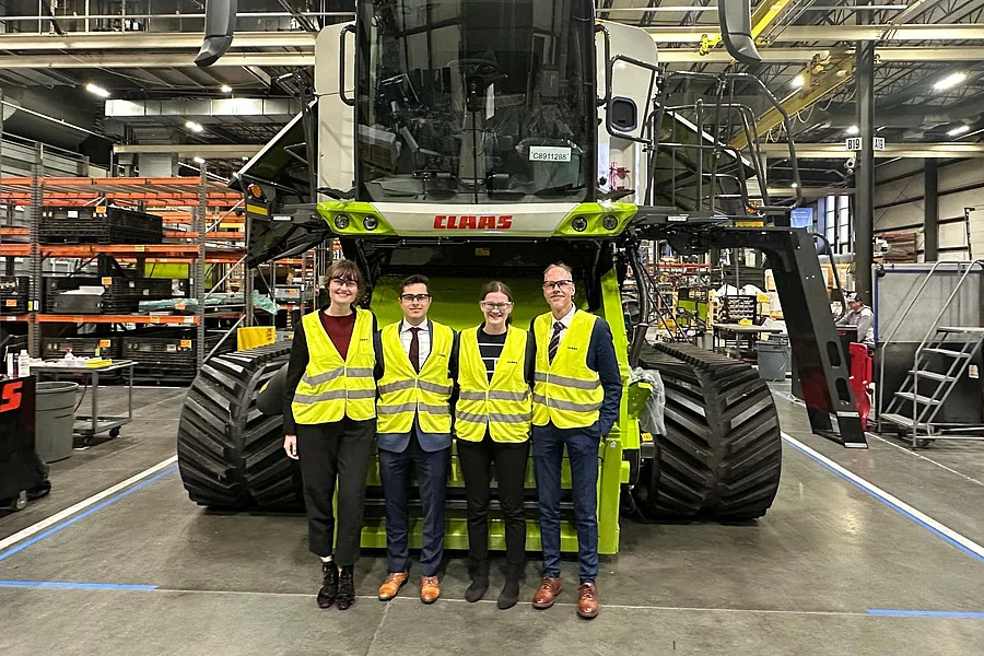 Four FES DC employees in yellow vests stand smiling in front of a large green Claas combine harvester inside a modern factory hall with shelves and equipment.