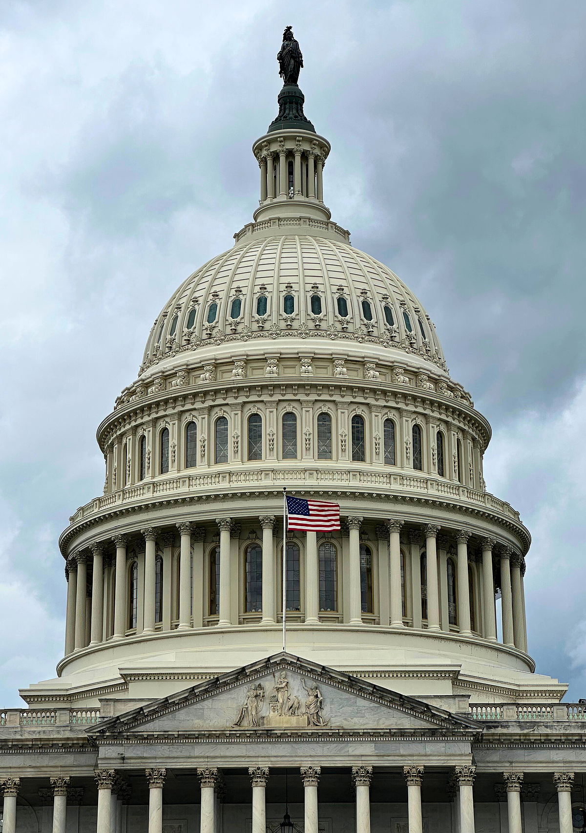 The white dome of the U.S. Capitol building with American flag, Statue of Freedom on top, against cloudy sky