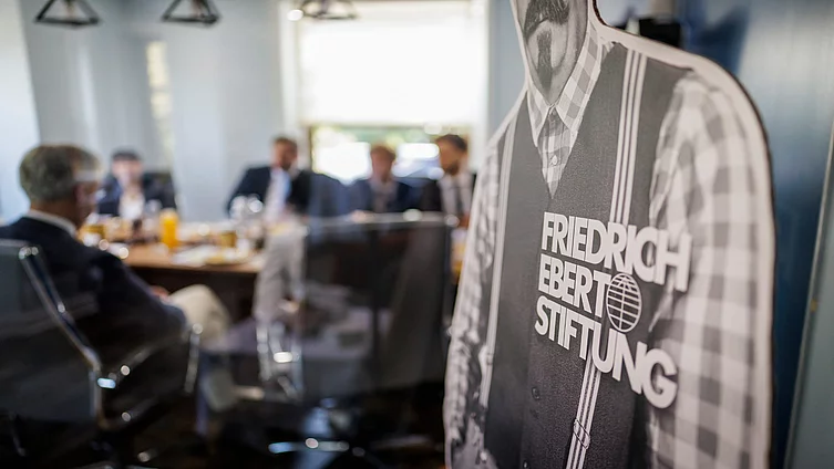 FES team and delegation seated around table with coffee cups during meeting, Friedrich Ebert Stiftung poster in foreground.