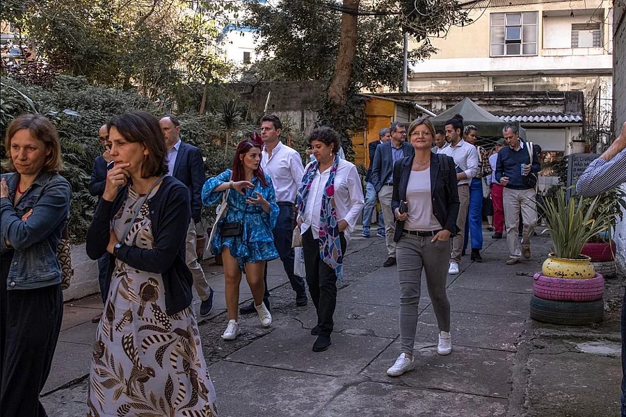 Group of 8 diverse professionals (4 women, 4 men) walking on urban outdoor path amid greenery and tents. Women in dresses, scarves, cardigans: men in suits, slacks. Smiling, chatting casually. Sunny day.