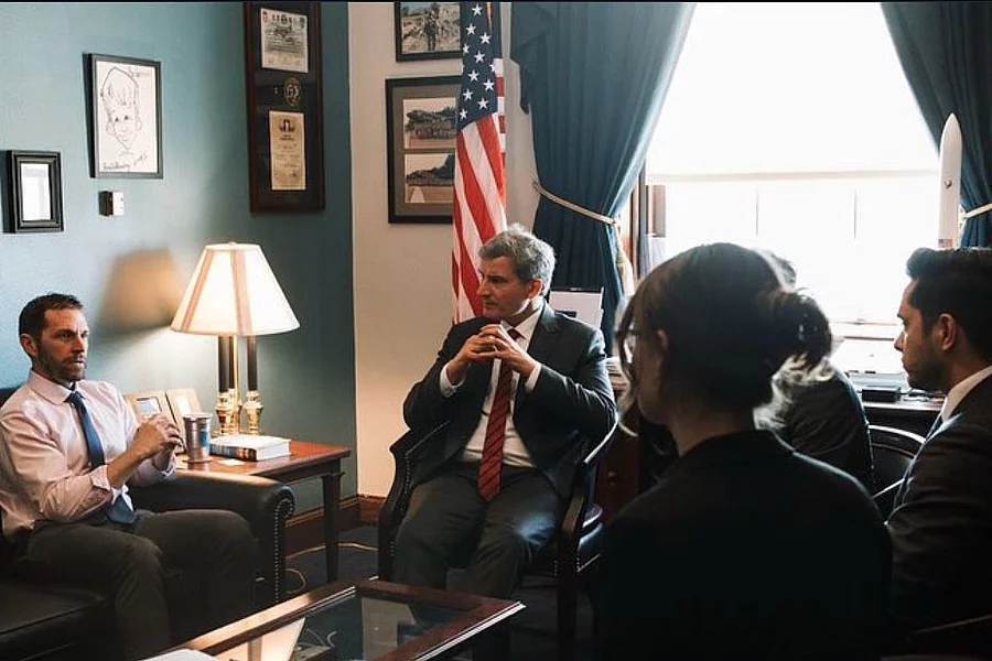 Office meeting: Right side shows FES staff listening to a Bundestag delegate who sitts in the middle next to a congressman to the left. In the background are teal walls, US flag, lamp, and window blinds.