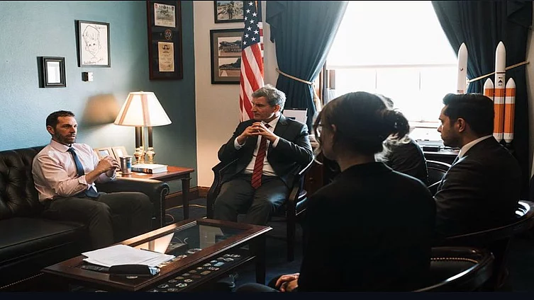 Office meeting: Right side shows FES staff listening to a Bundestag delegate who sitts in the middle next to a congressman to the left. In the background are teal walls, US flag, lamp, and window blinds.